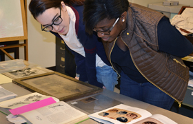 Two women lean over a table to examine books and framed photos.